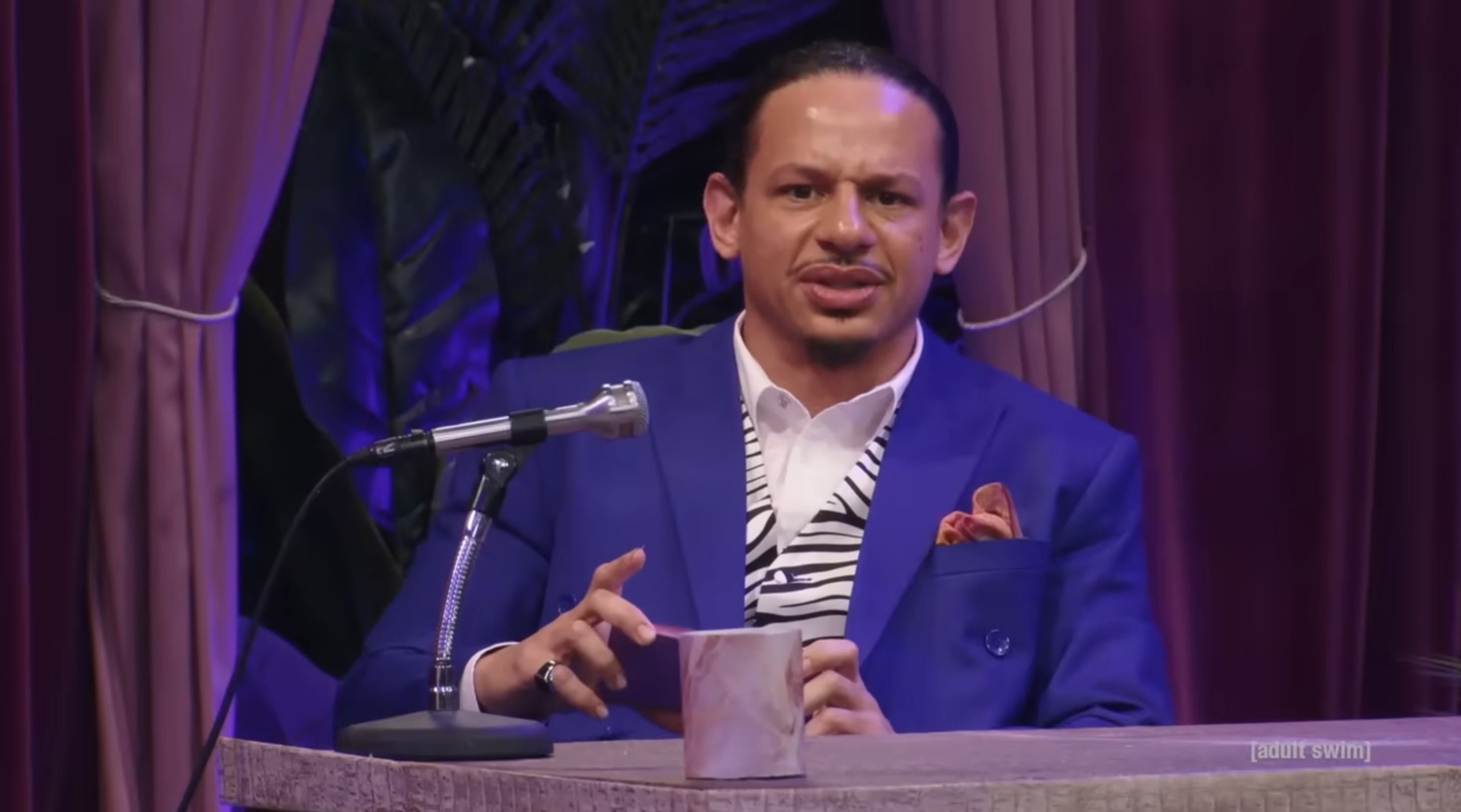 Andre seated at a desk with a microphone, wearing a blue suit and a zebra-striped shirt, appearing to speak or perform on a set