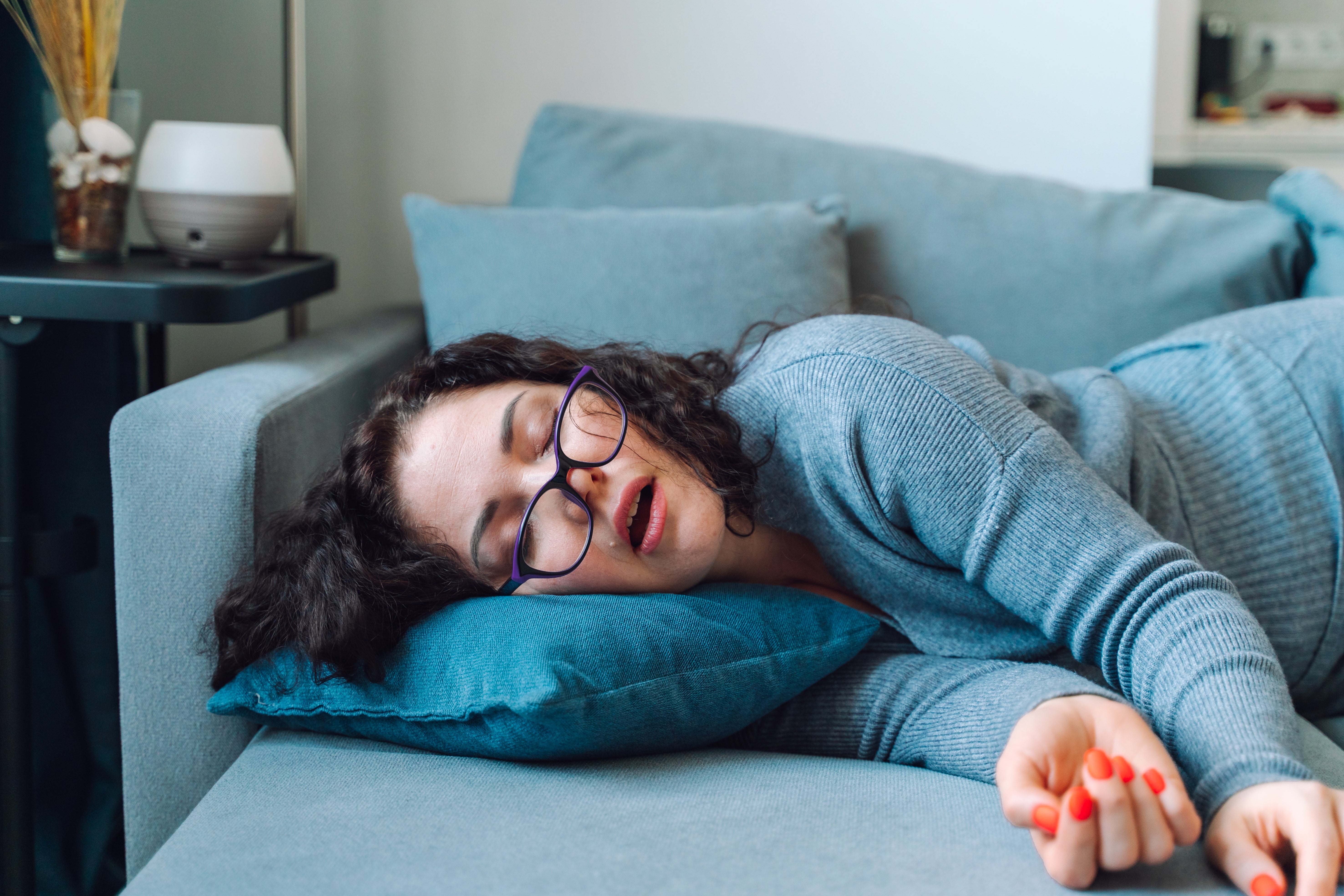 Person with glasses asleep on a couch, wearing a cozy sweater, resting on a cushion