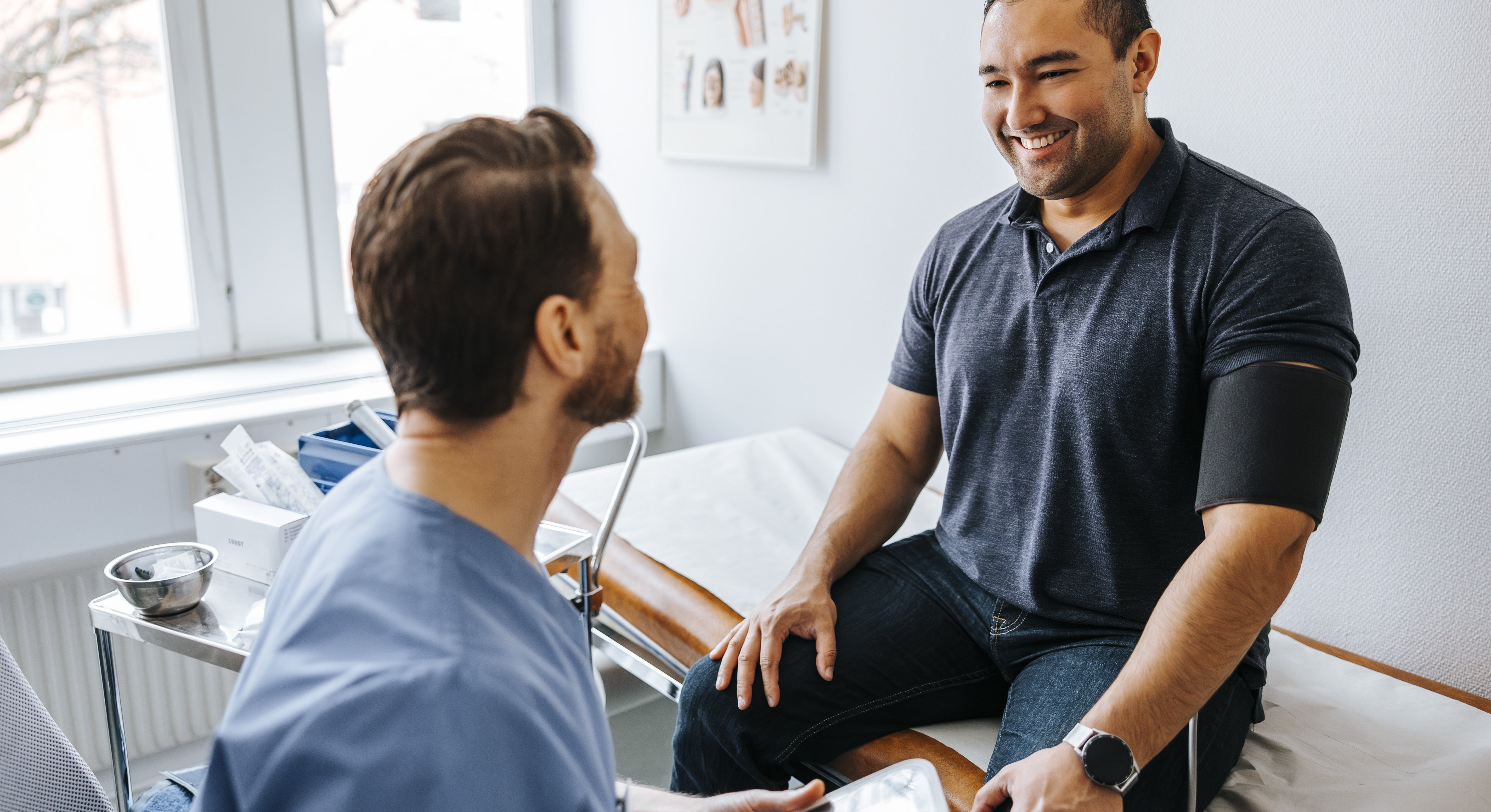 Doctor in scrubs checks a patient's blood pressure. The patient smiles, sitting on a medical bench in a clinic room