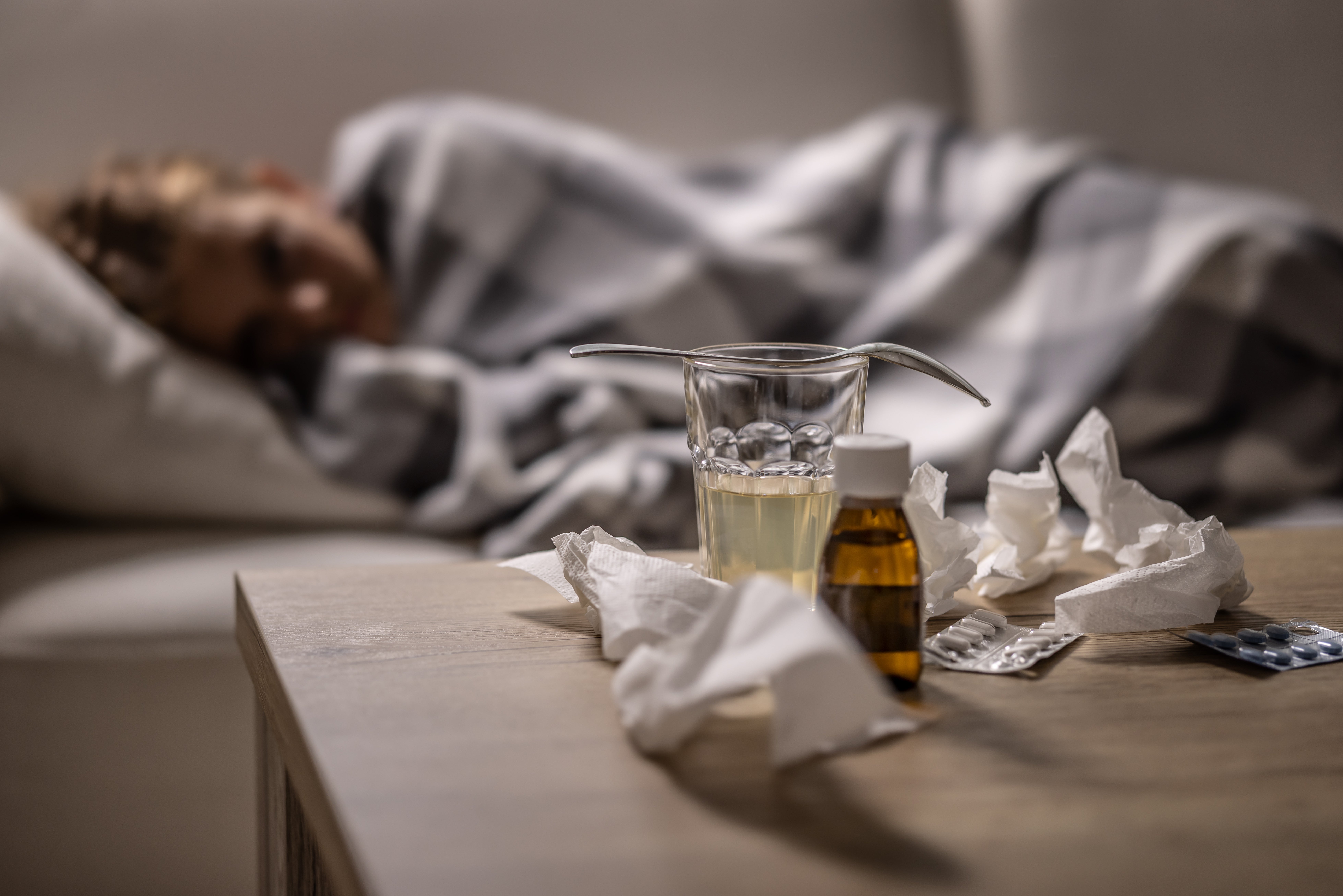Person resting under a blanket on a couch, with medicine, a glass of liquid, and tissues on a nearby table