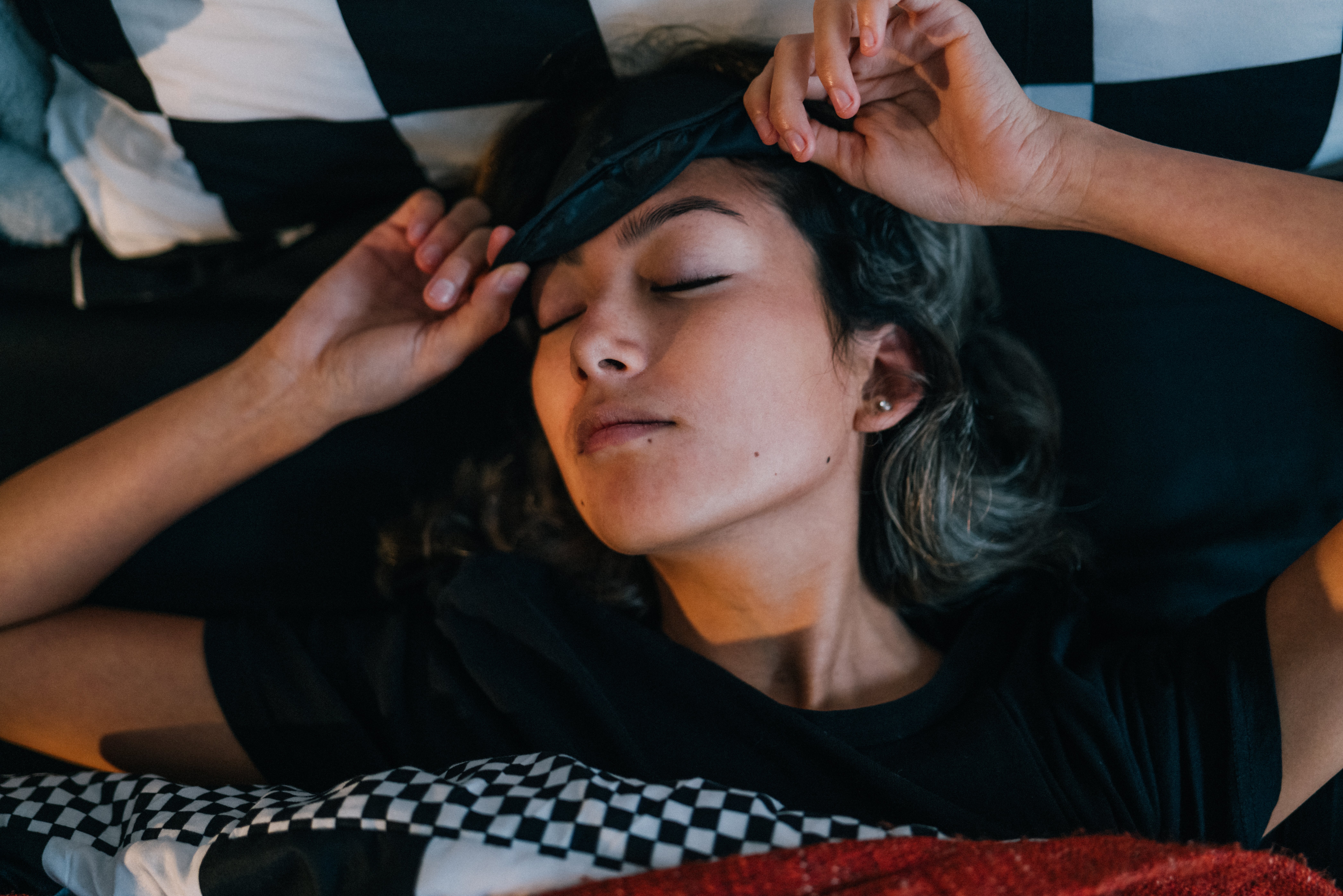 Person lying in bed, eyes closed, gently adjusting an eye mask, appearing relaxed or asleep