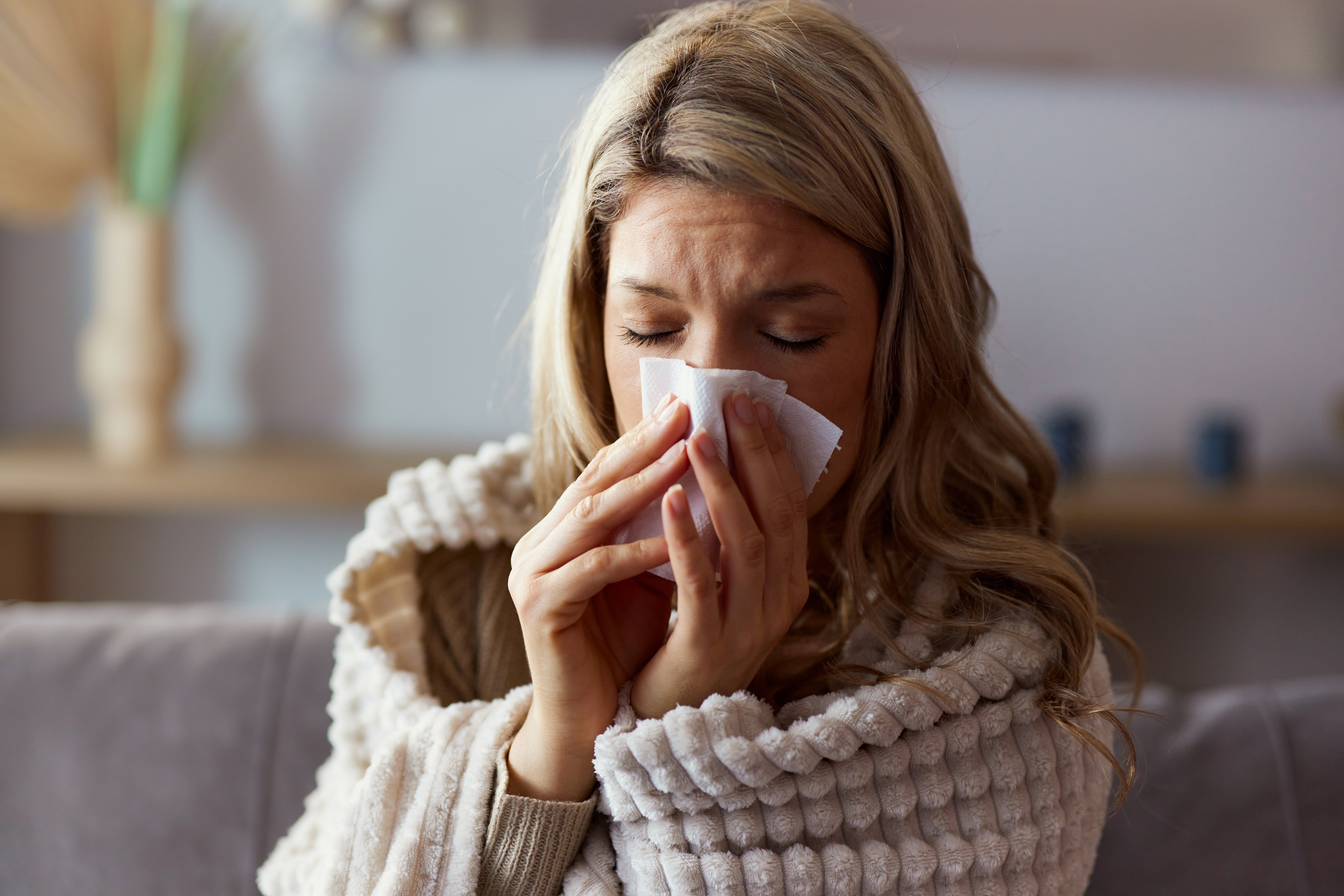 A woman sitting on a couch, wrapped in a textured blanket, blowing her nose with a tissue