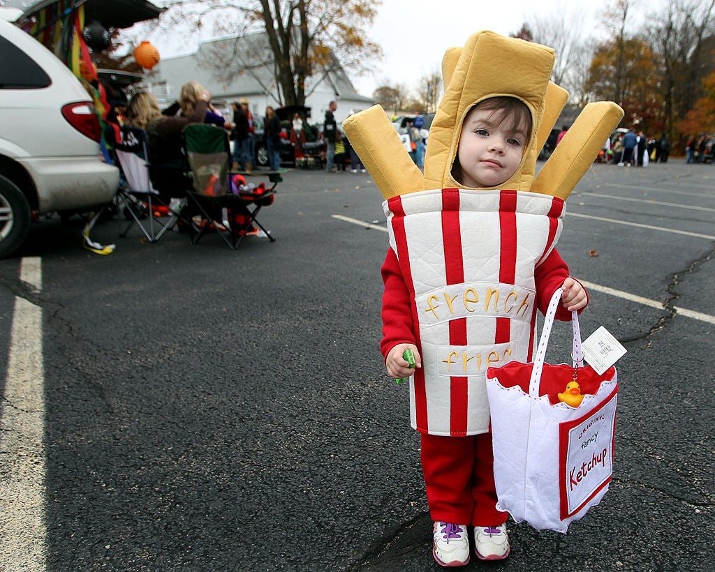 Child wearing a French fries costume, holding a bag labeled "ketchup" at an outdoor event with parked cars and people in the background