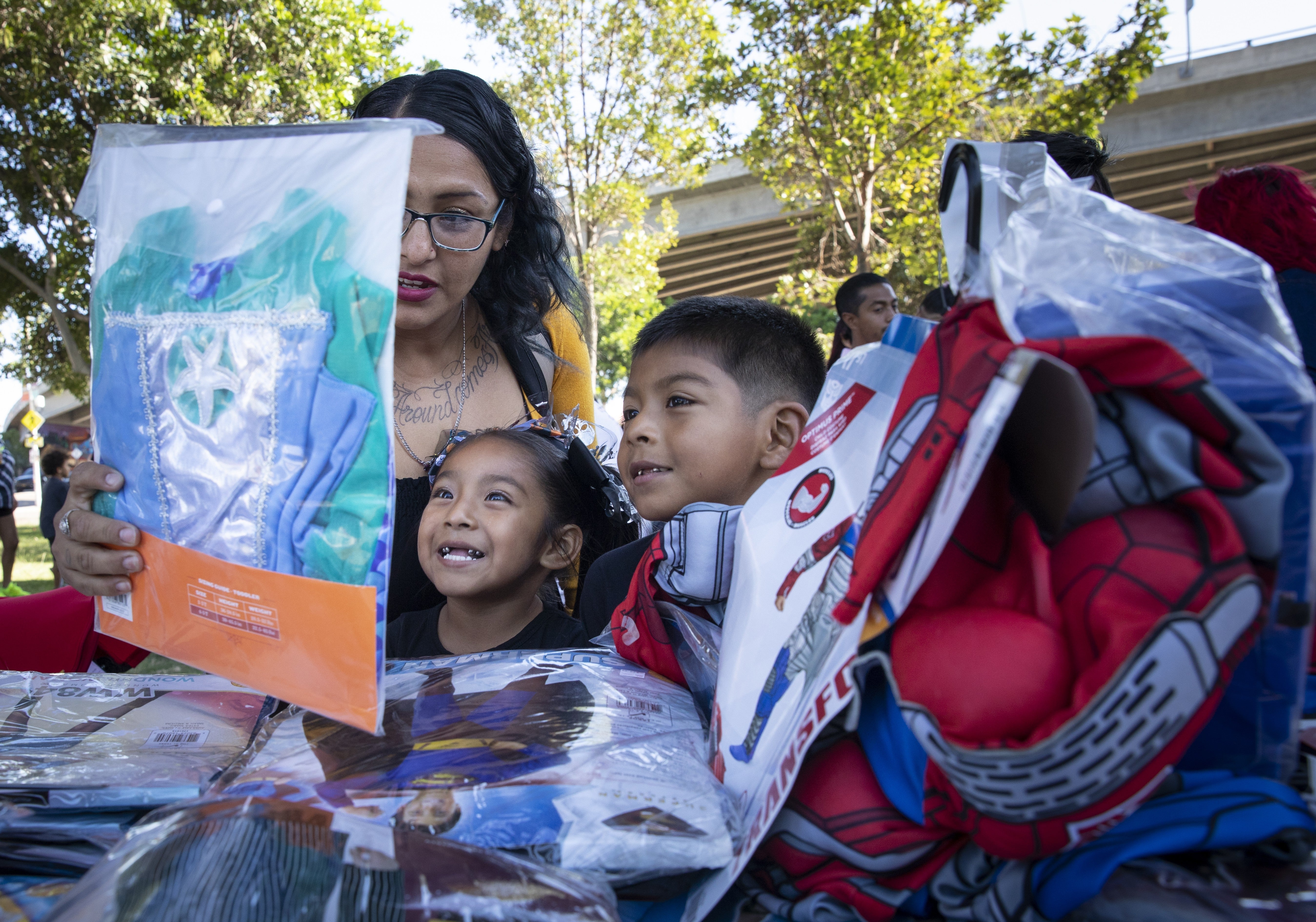 A family happily looks at superhero costumes at an outdoor event, surrounded by other attendees and trees in the background