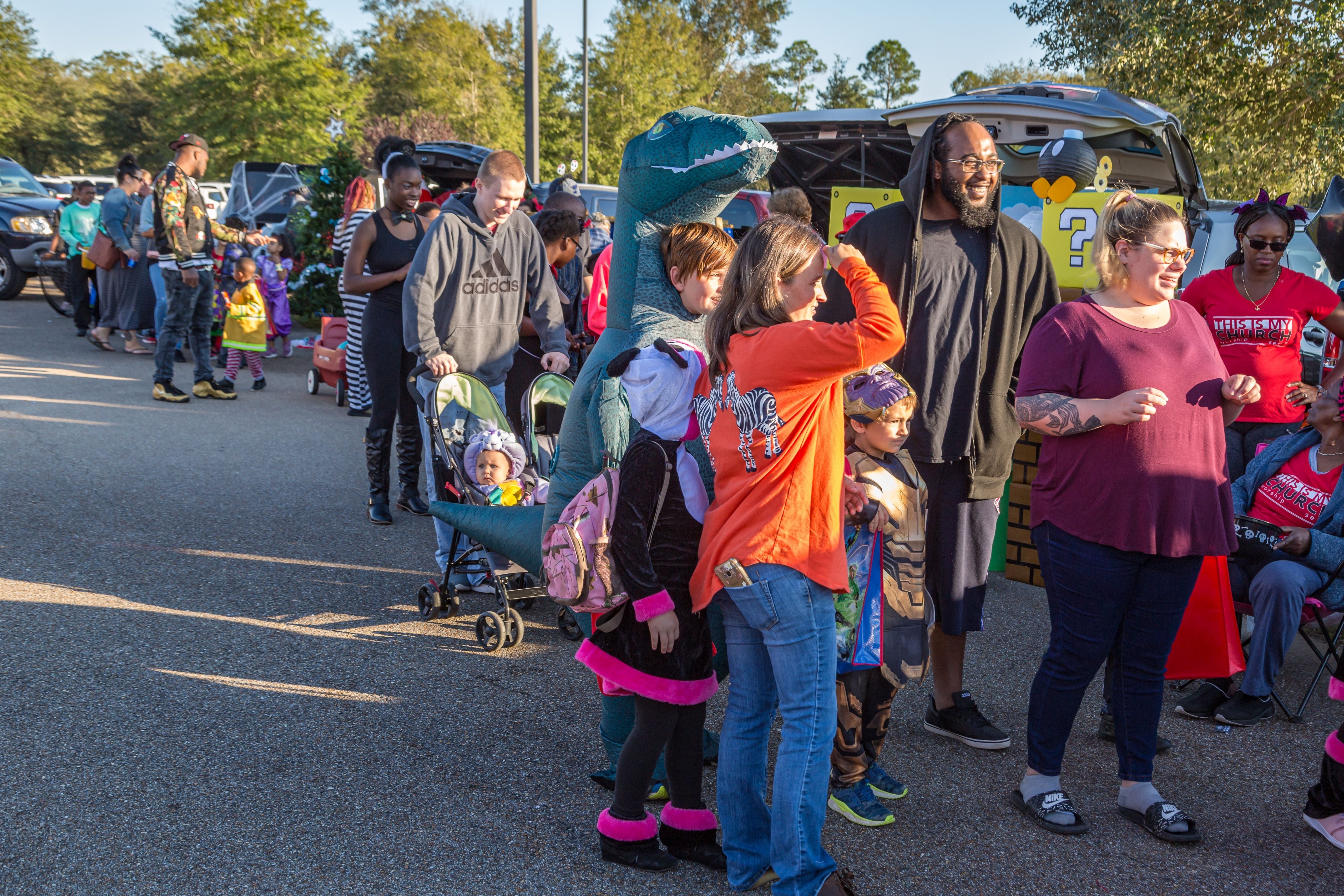People in costumes, including a dinosaur suit, line up outdoors near decorated cars, possibly at a festival or fair event