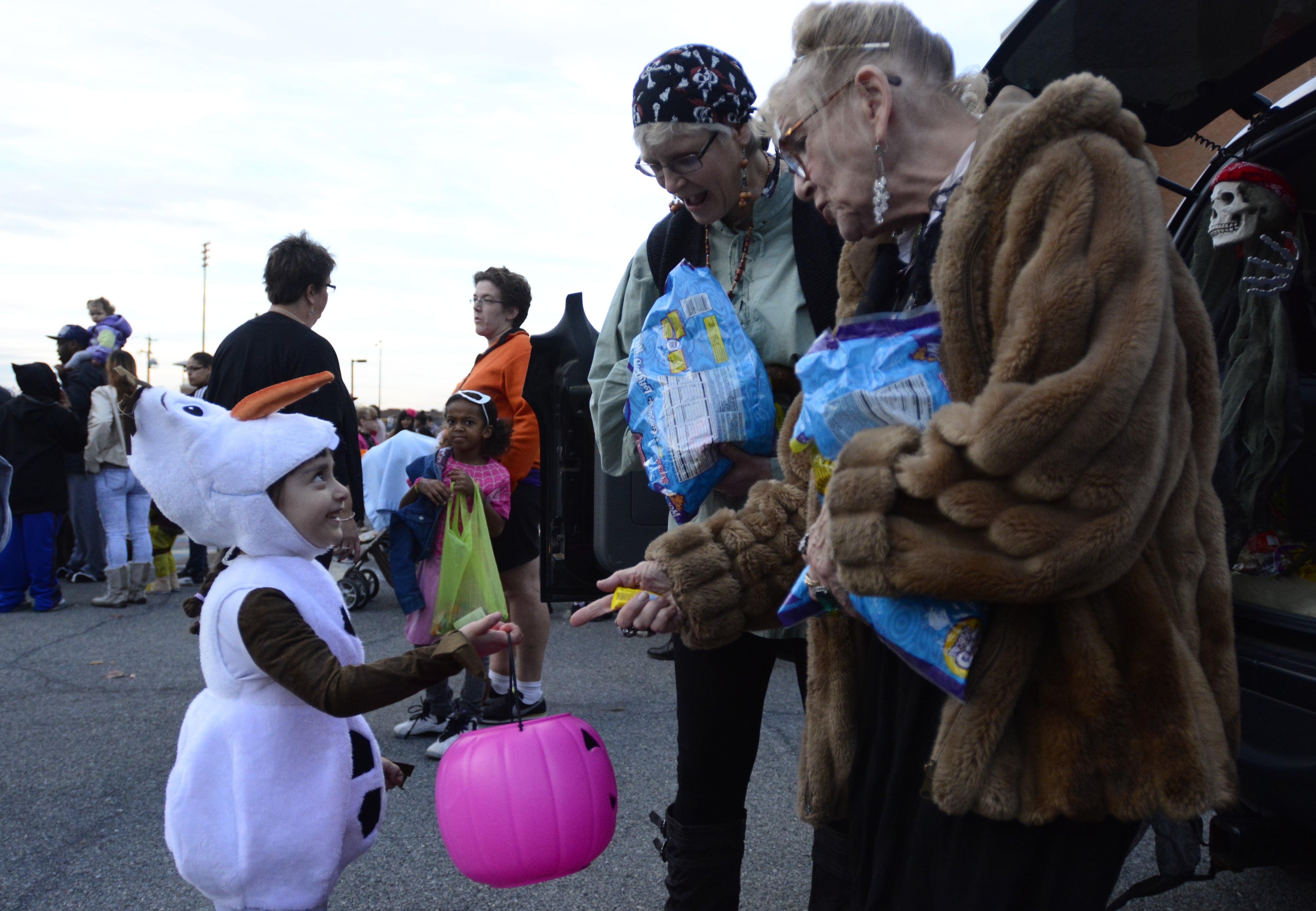 Child in a snowman costume trick-or-treating, receiving candy from two older adults holding bags at a community event