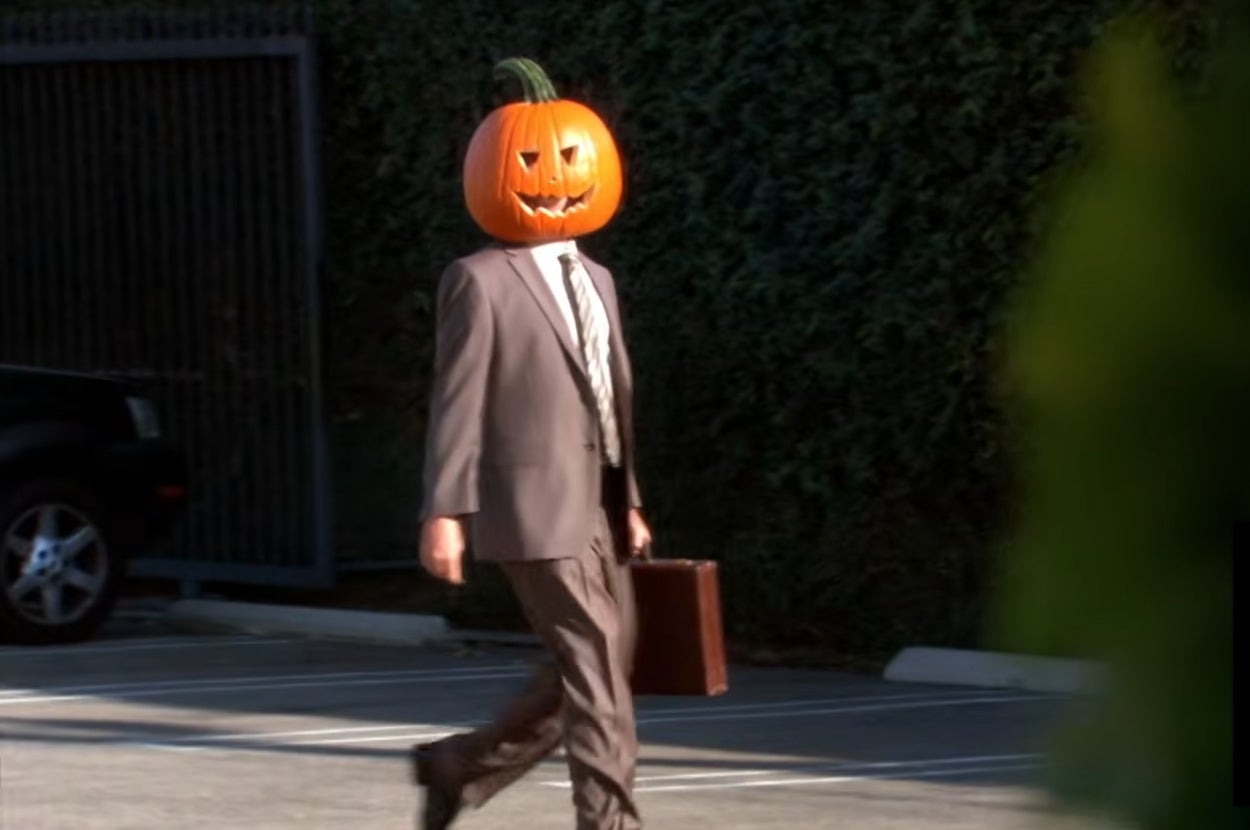 Dwight from "The Office" in a suit walking in a parking lot with a carved pumpkin on his head, holding a briefcase