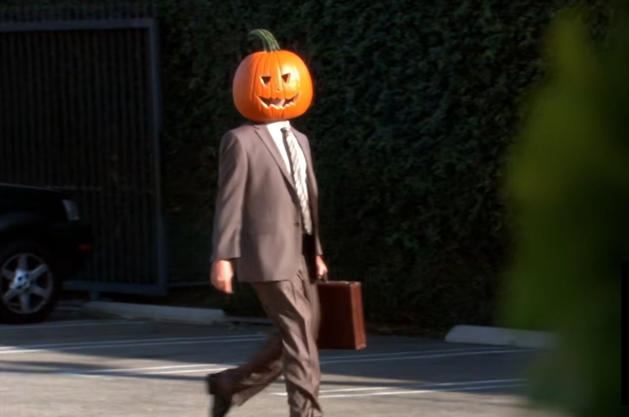 Dwight from "The Office" in a suit walking in a parking lot with a carved pumpkin on his head, holding a briefcase