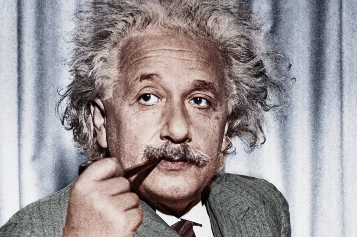 A person with distinct white hair and a mustache holds a pipe, wearing a suit, in front of a plain backdrop
