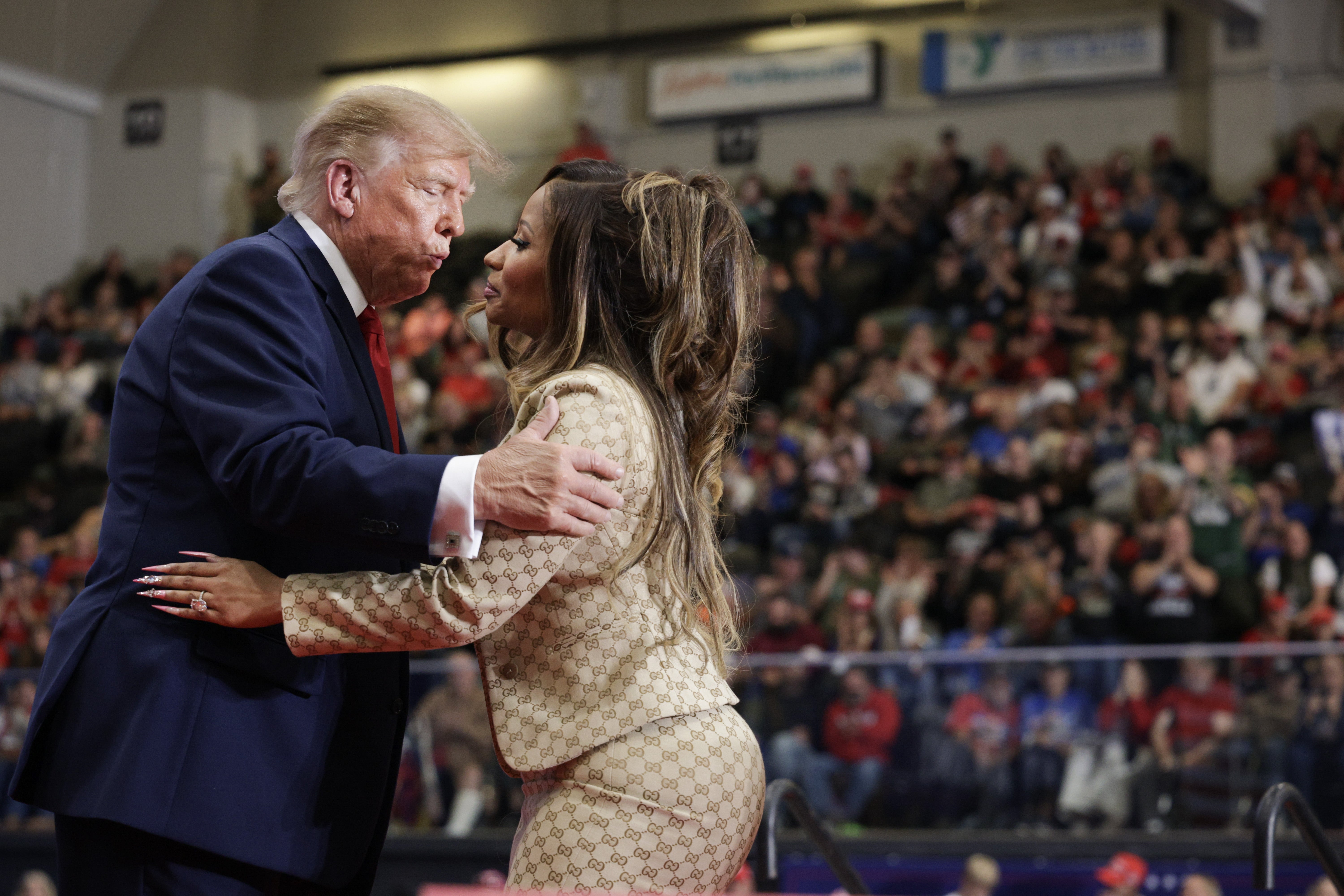 A man in a suit greets a woman in a patterned outfit at a public event, in front of a large crowd