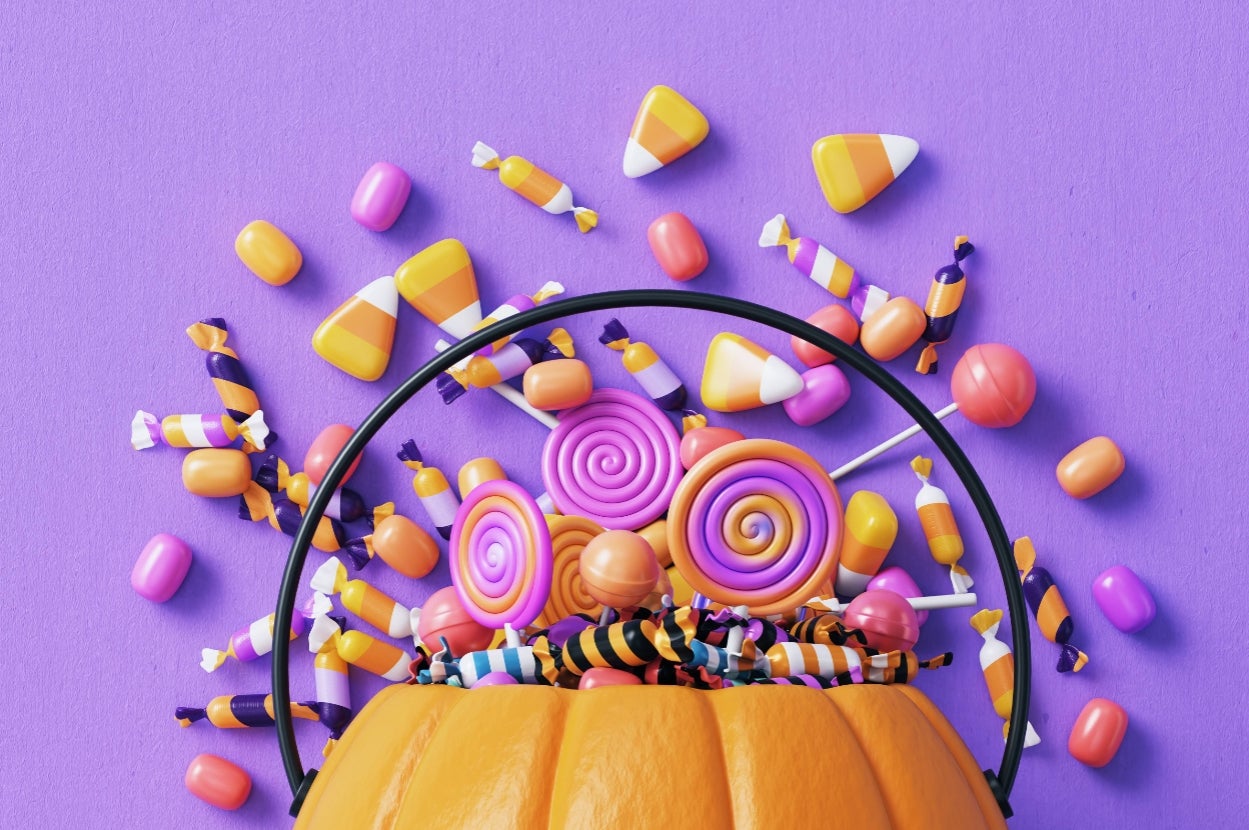 A pumpkin bucket filled with colorful lollipops, candies, and candy corn, arranged artfully against a plain background