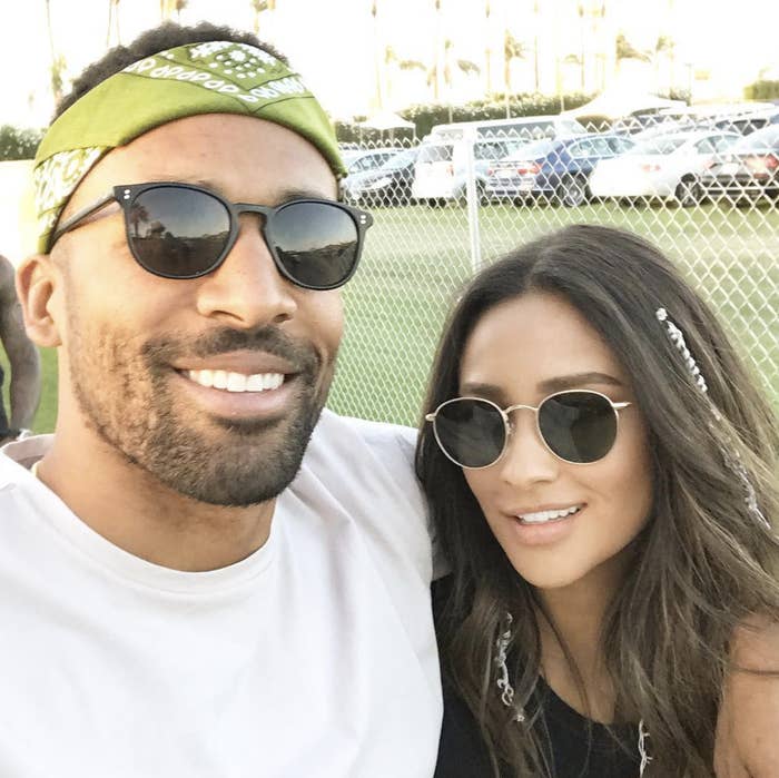 Two people wearing sunglasses smiling for a selfie outdoors, with festival elements in the background