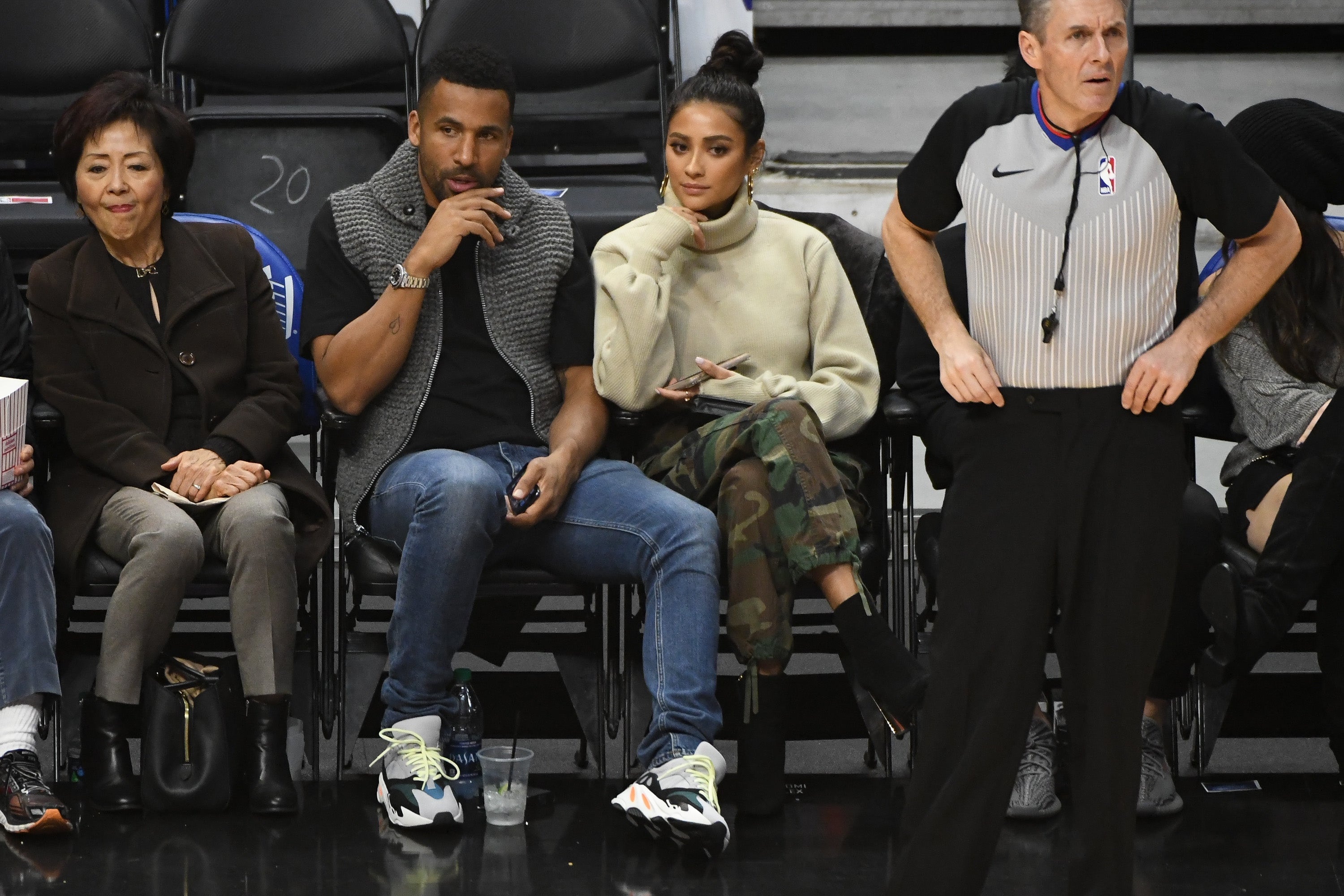 People sitting courtside at a basketball game, including a woman in a turtleneck and camo pants, and a man in a patterned sweater. An official stands nearby