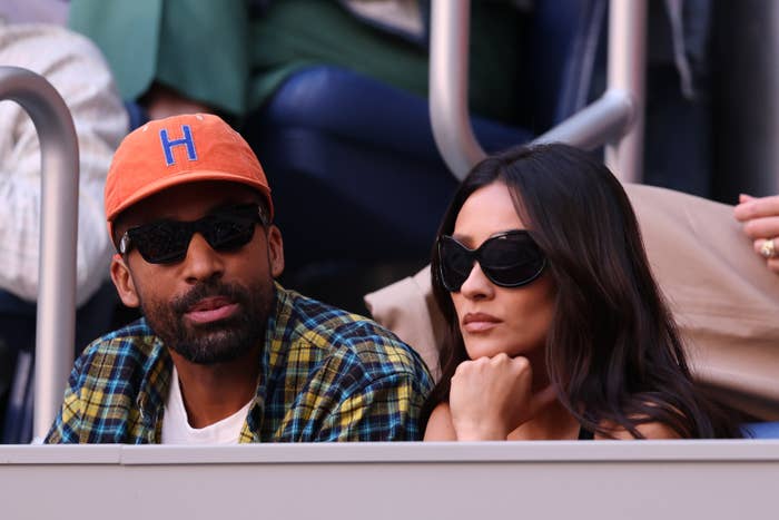 Two people sitting together in an outdoor setting, both wearing sunglasses. One wears a plaid shirt and an orange cap with an "H" on it