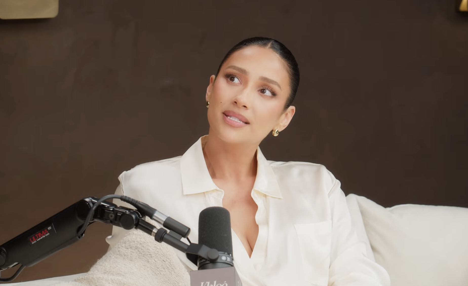 A person in a white blouse sits on a sofa, speaking into a microphone during an interview