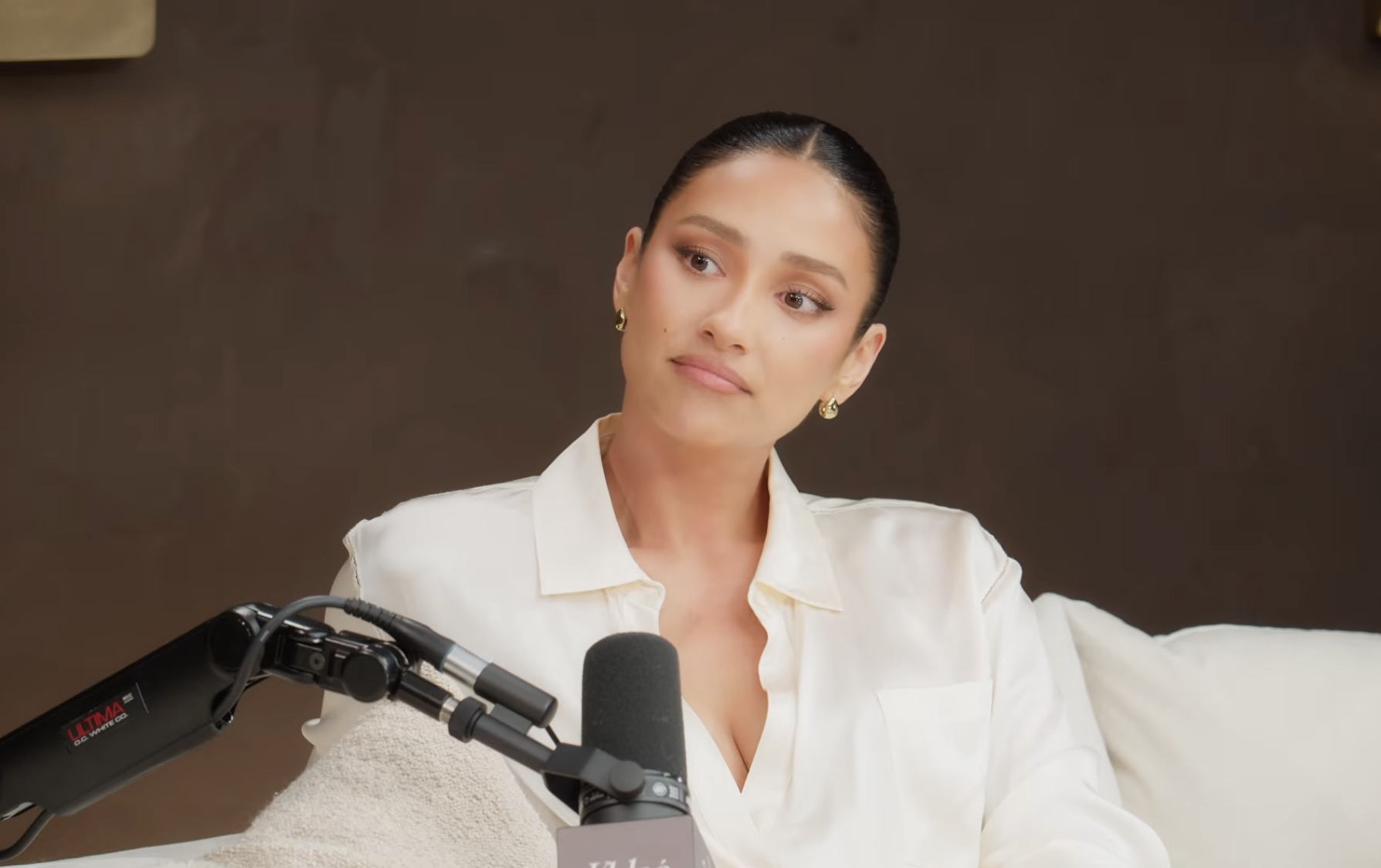 Person in a white blouse, sitting with a microphone, appearing engaged in a conversation