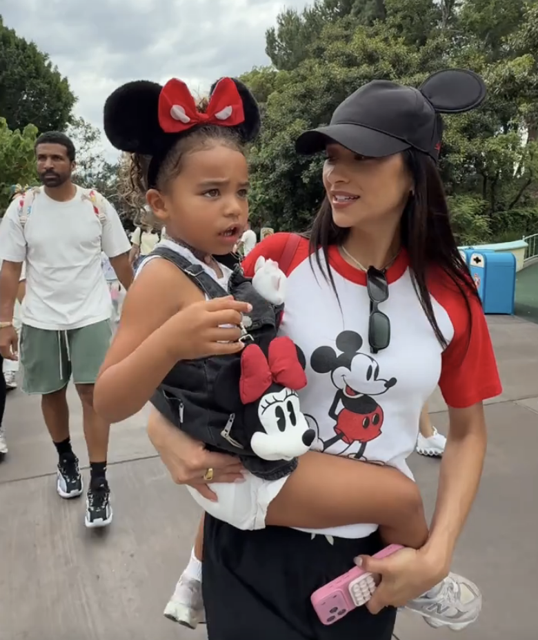 A woman in a Mickey Mouse shirt and cap holds a child wearing Minnie Mouse ears and backpack at a theme park