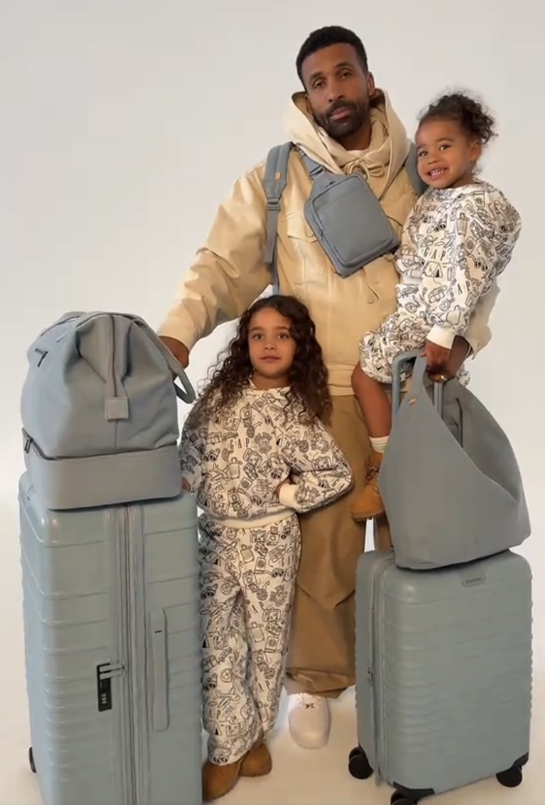 Man with two young children pose with matching gray luggage; children wear matching printed outfits; man wears casual hoodie and pants
