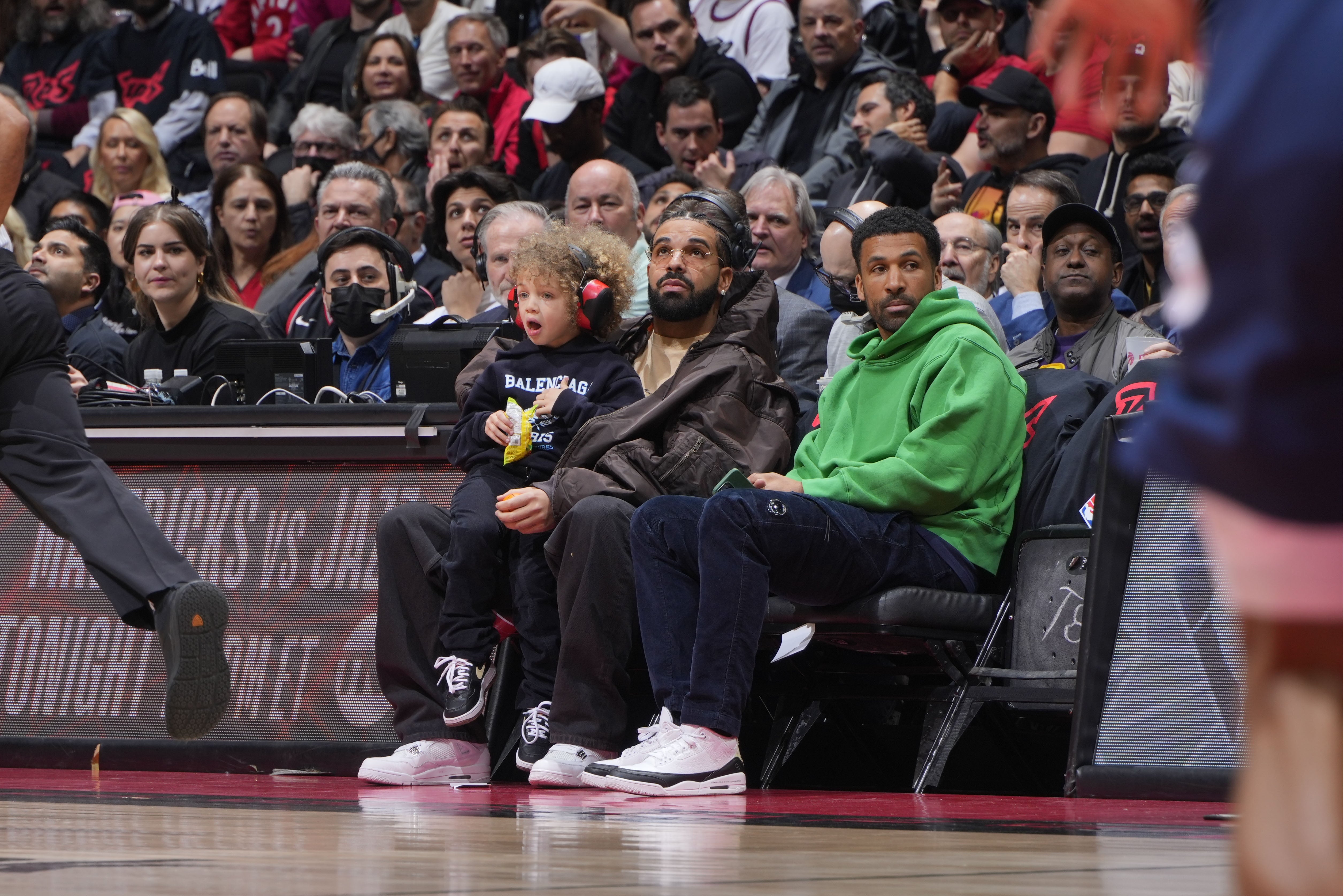 A crowd watches a basketball game. Two adults and a child with headphones sit courtside, focusing on the action