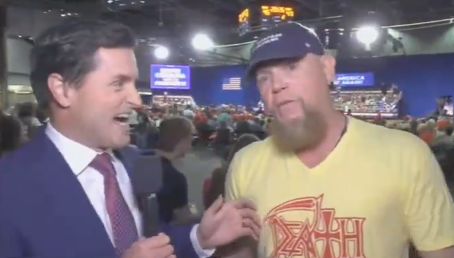 Two men talking at an indoor event. One in a suit holds a microphone, the other in a casual T-shirt and cap