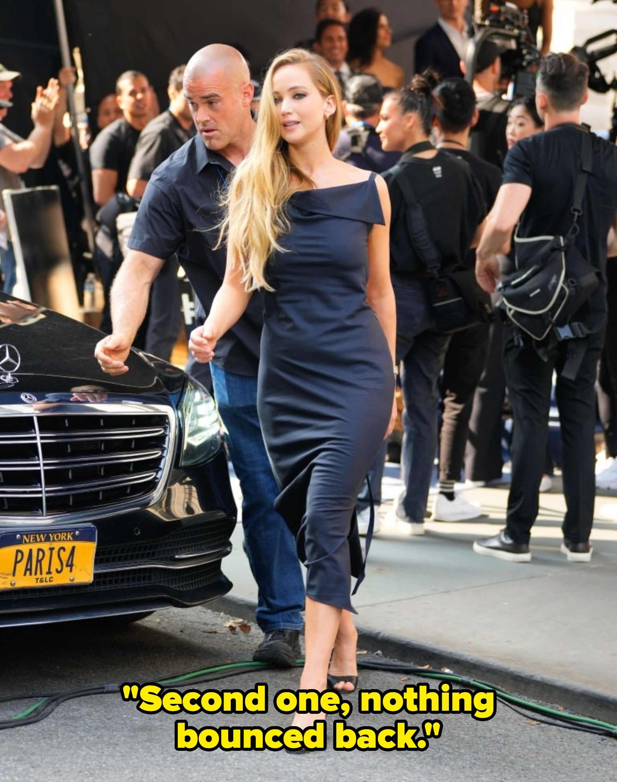 A woman in a sleek, asymmetrical dress walks confidently past a car on a city sidewalk, escorted by a security guard. The scene is lively with onlookers