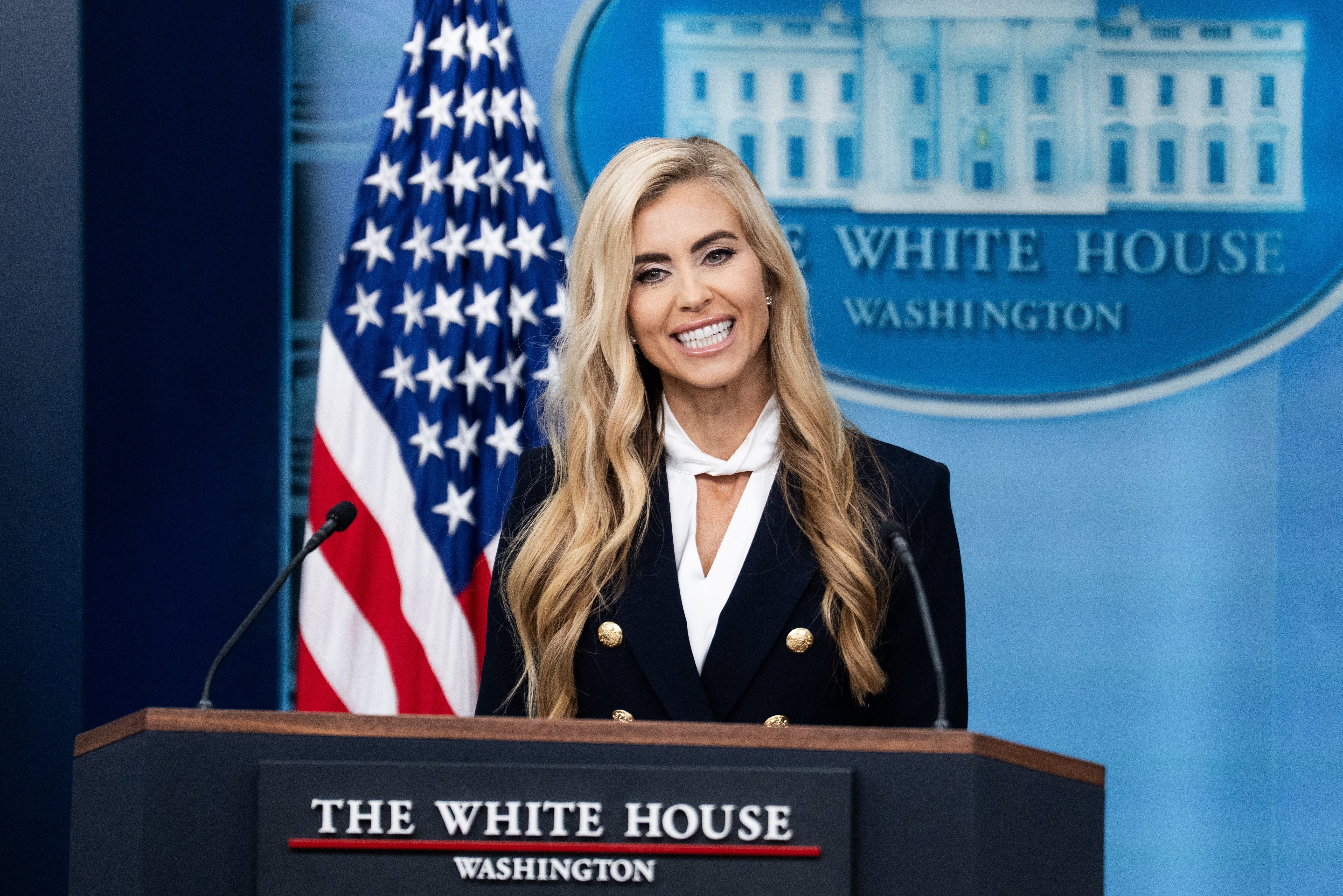 Woman at a podium in formal attire, speaking at a press briefing with a White House backdrop