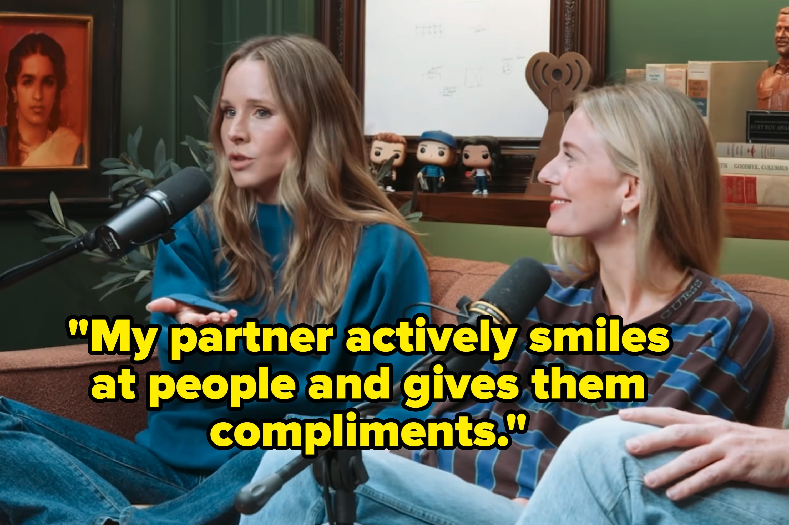 Kristen Bell and Justine Lupe sitting on a couch, each speaking into a microphone during a podcast recording. Books and framed art are in the background