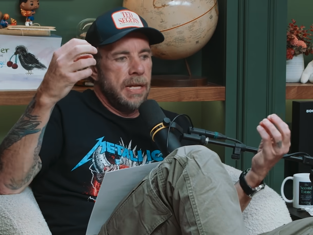 Dax Shepard in a casual setting, wearing a cap and graphic T-shirt, gesturing while speaking into a microphone during a podcast or interview
