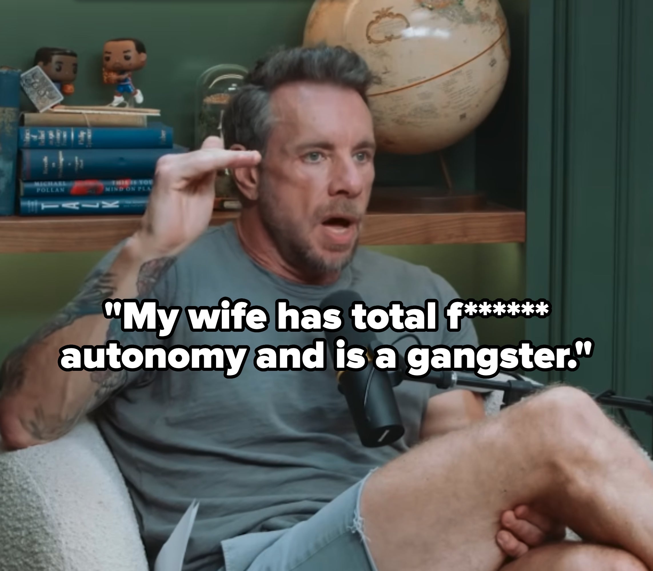 Dax Shepard with tattoos wearing a casual t-shirt and shorts, speaking into a microphone in a room with books and a globe on a shelf