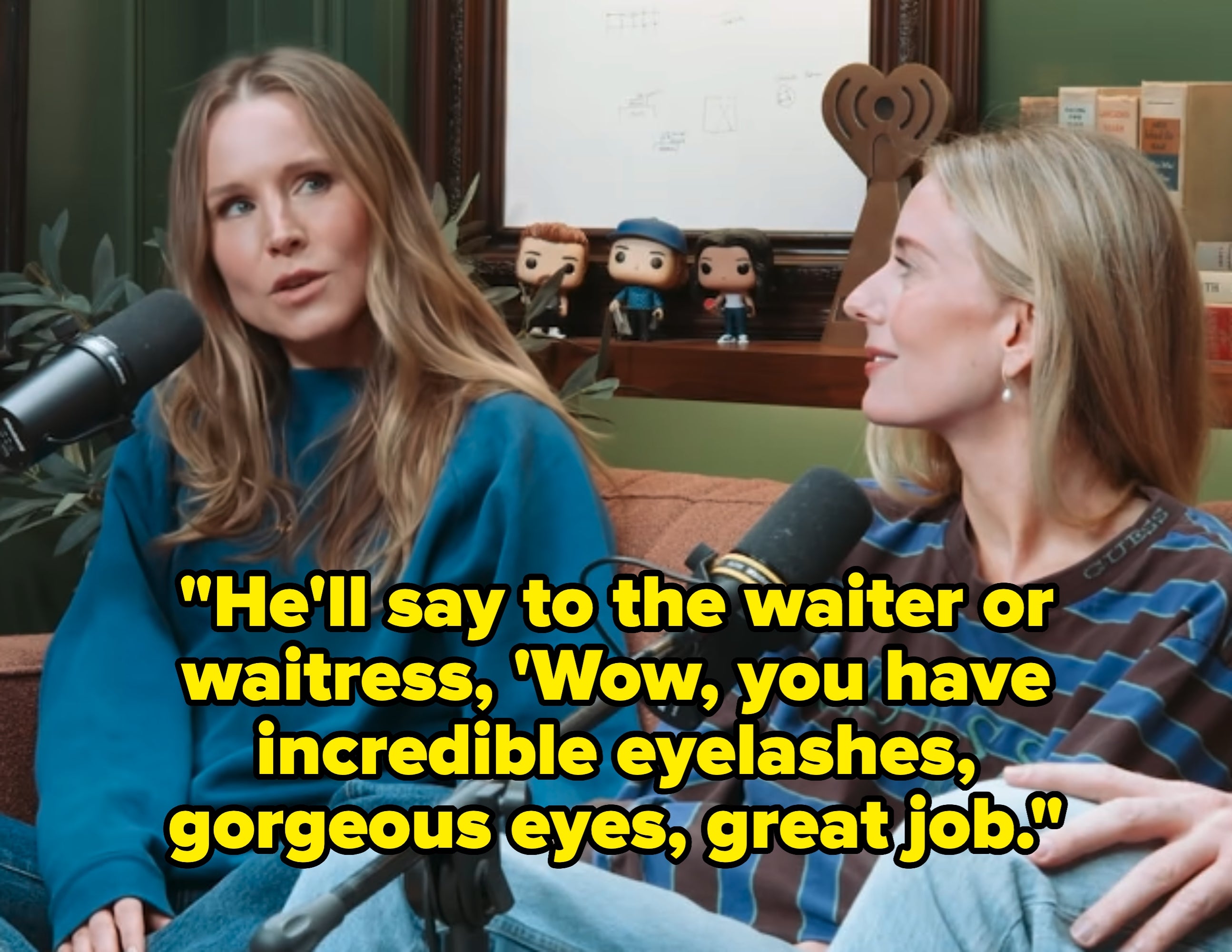 Kristen Bell and Justine Lupe sitting on a couch in a podcast setting, both speaking into microphones, surrounded by pop culture decor