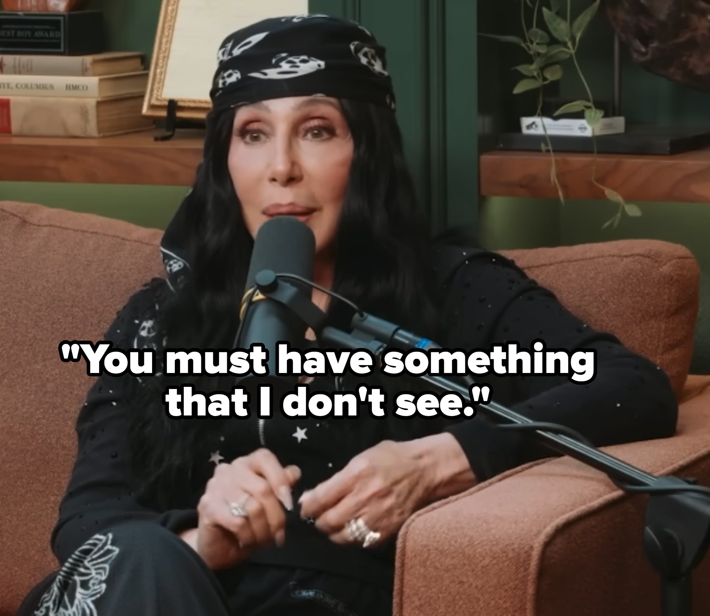 Cher with long dark hair wearing a bandana and speaking into a microphone while seated on a couch
