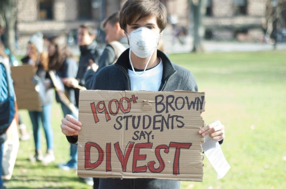 A Brown University student wearing a face mask holds up a sign supporting the student-led divestment campaign.