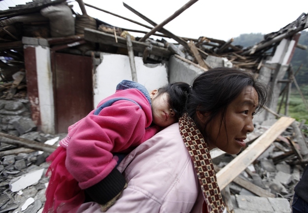 A mother carrying her sleeping daughter walks past debris in a mountainside village.