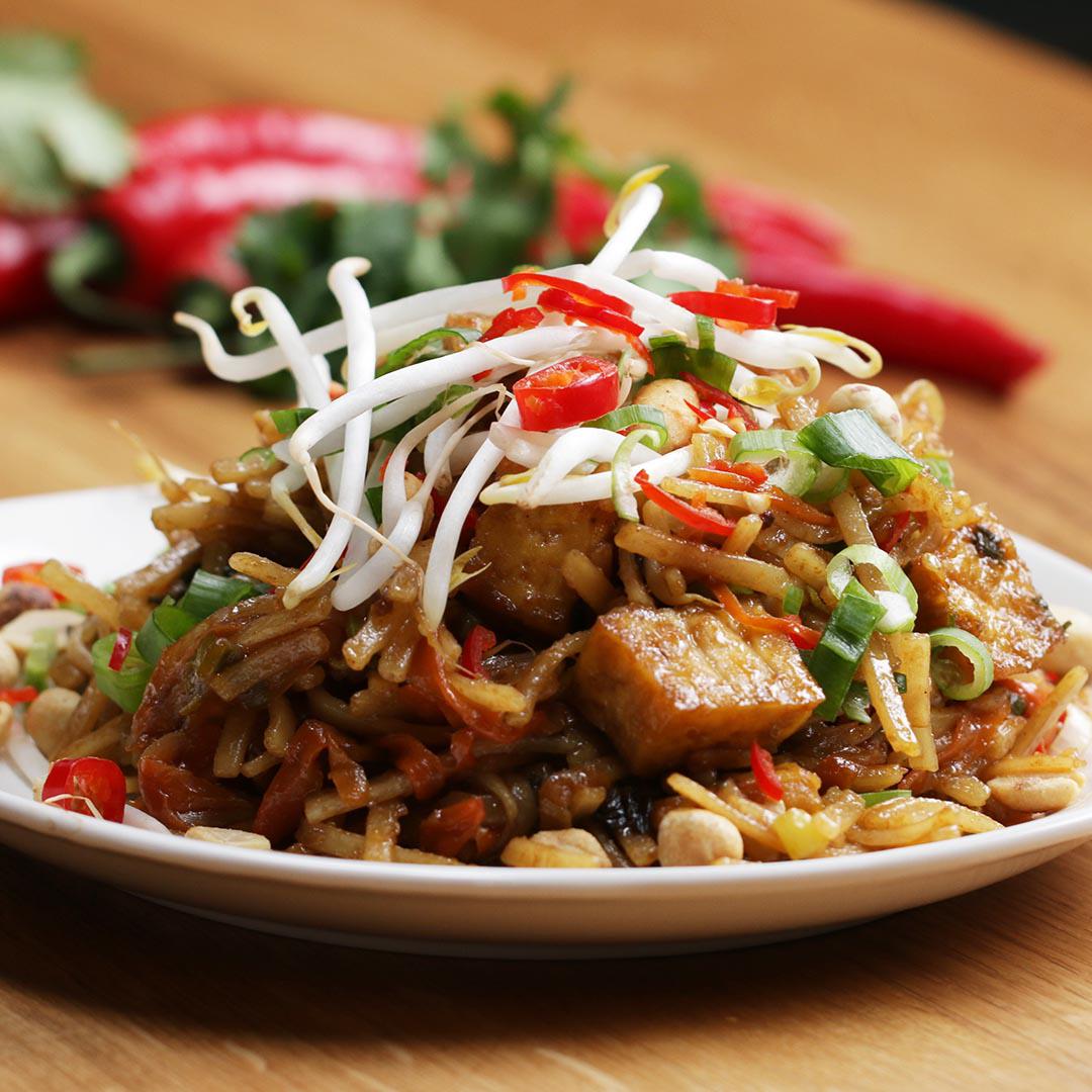 A plate of stir-fried noodles topped with tofu, bean sprouts, chopped chili, and green onions, served on a wooden table with fresh peppers in the background