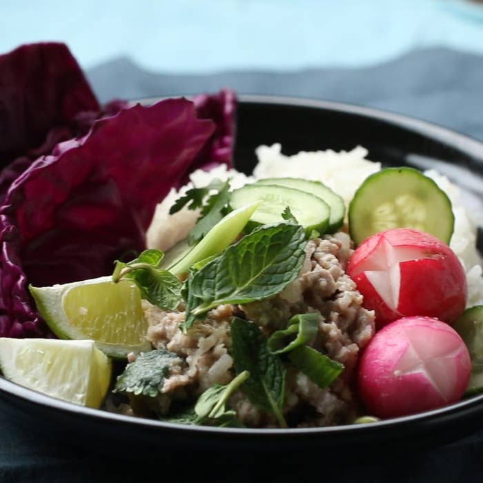 A bowl of minced meat with fresh mint, sliced cucumbers, radishes, lime wedges, red cabbage, and rice, arranged attractively