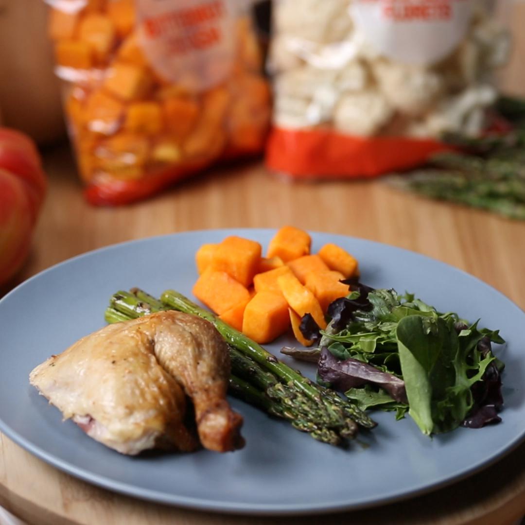 A plate with roasted chicken, asparagus, cubed sweet potatoes, and mixed greens. Bags of vegetables in the background