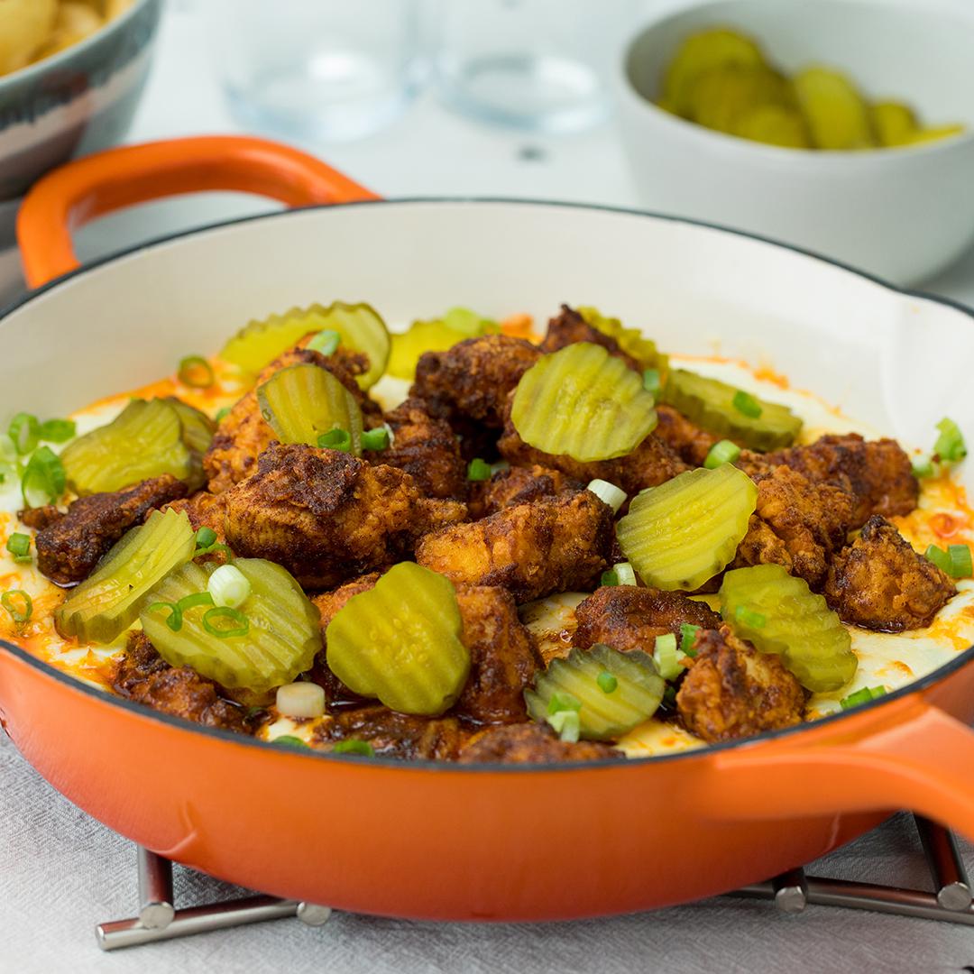 recipes A dish of fried chicken pieces topped with pickles and green onions in a pan, served with a bowl of pickles in the background