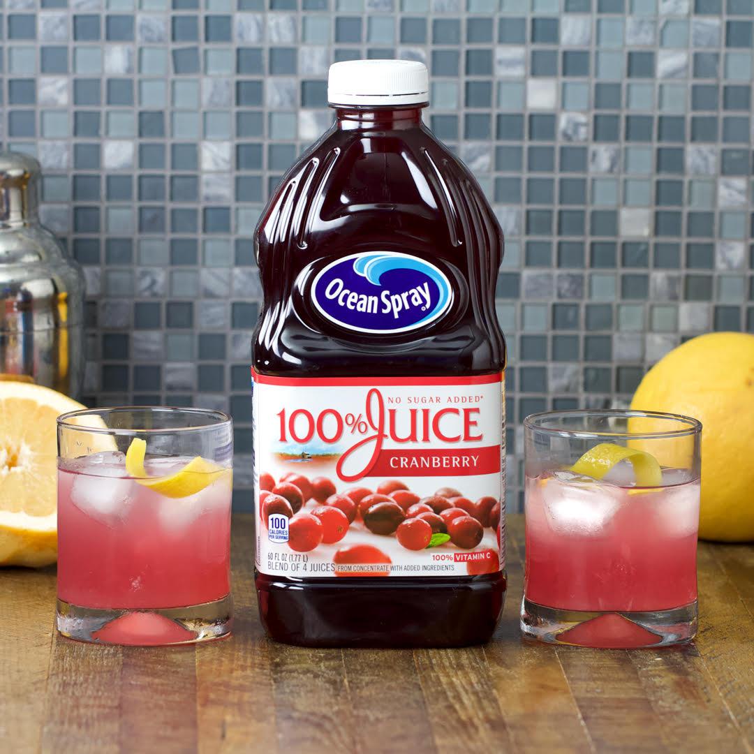 Ocean Spray 100% Cranberry Juice bottle with two glasses of pink juice, garnished with lemon slices, displayed on a counter with fruits