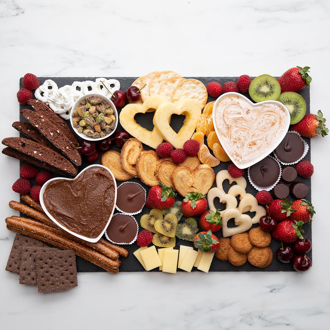 A heart-themed charcuterie board with assorted cheeses, fruits, nuts, chocolate, pretzels, and bread on a slate serving tray