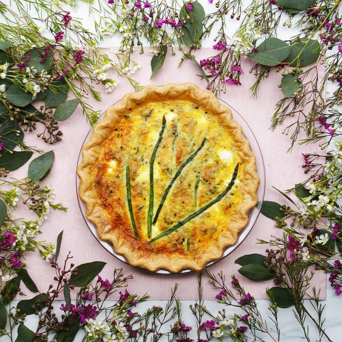 A baked quiche garnished with asparagus, surrounded by decorative flowers and greenery connected a pinkish cloth