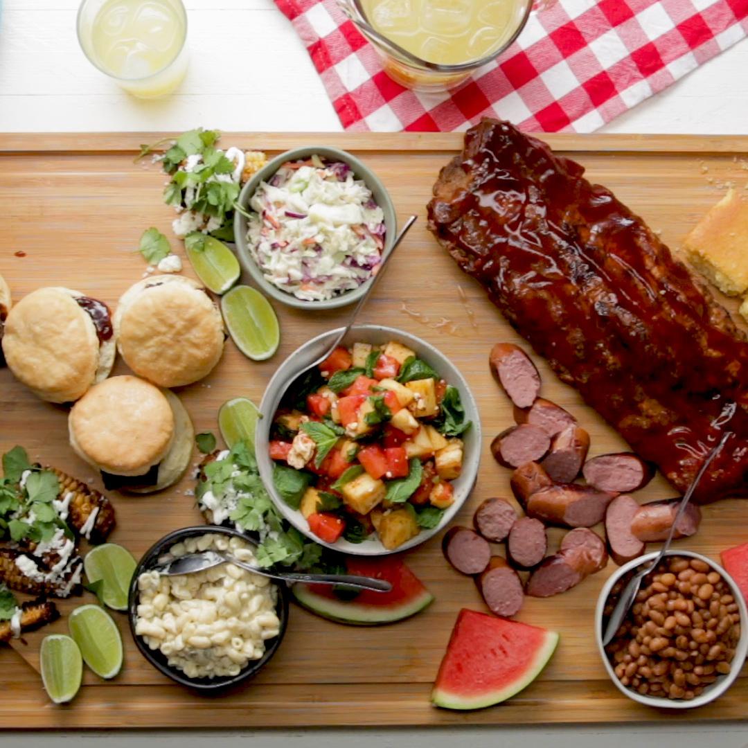 A wooden board with BBQ ribs, sausage, sliders, watermelon, mac &#x27;n&#x27; cheese, coleslaw, corn, and baked beans arranged for a summer meal