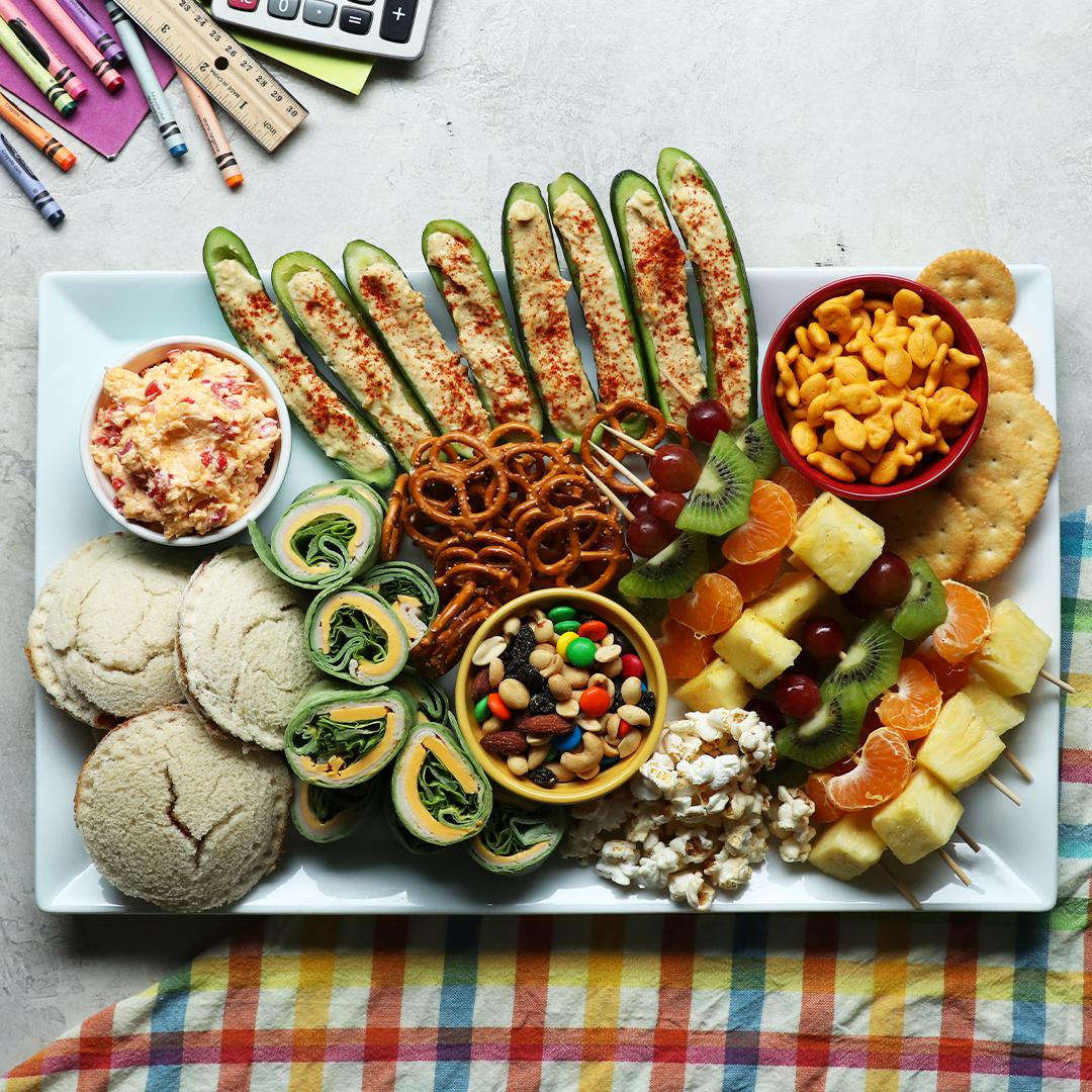 A platter with assorted snacks: cucumber bites, cheese crackers, sandwiches, wraps, fruit skewers, mixed nuts, and popcorn, arranged invitingly