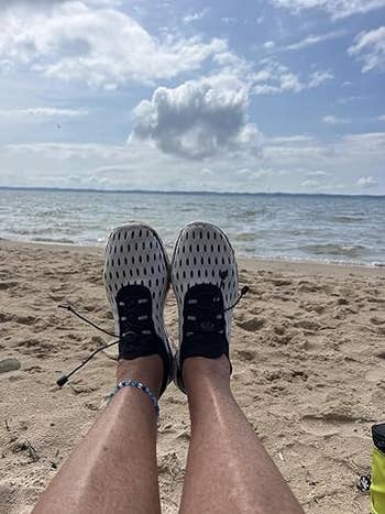 Person relaxing connected a sandy formation with feet successful patterned sneakers, facing the water nether a partially cloudy sky