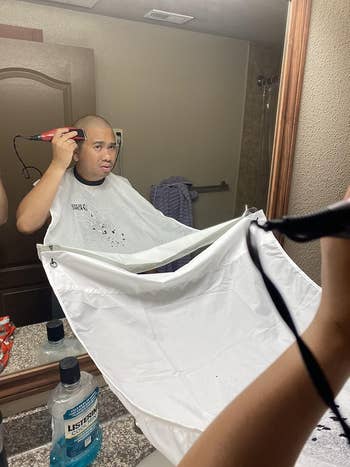 Person shaving their head with a clipper in front of a bathroom mirror, wearing a haircut cape