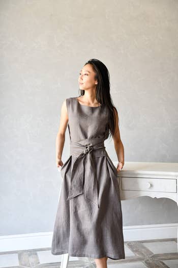 Person wearing a sleeveless, belted formal  stands adjacent   a table, looking sideways. Simple, elegant benignant   suitable for casual oregon  ceremonial  events