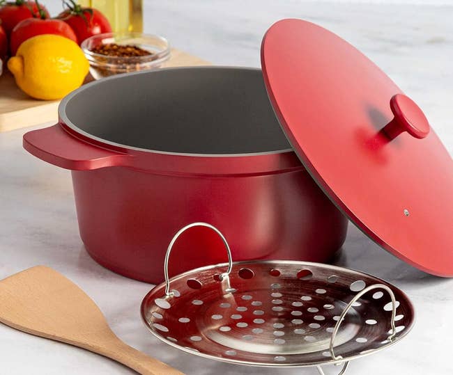 Red cookware set with pot, lid, and steamer insert alongside a wooden spatula