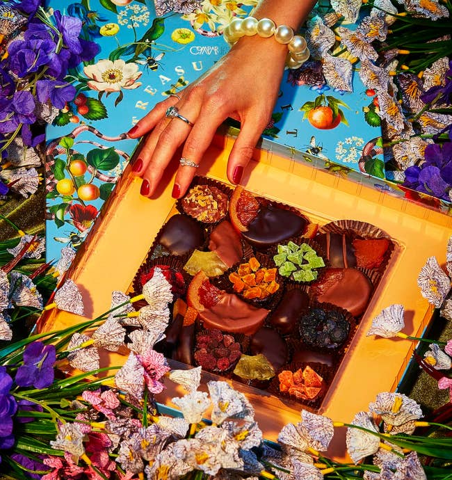 a model pulling a chocolate out of the opened gift box surrounded by flowers 