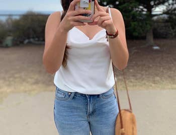 Reviewer holding phone taking selfie in white cowl neck top and denim jeans, holding straw bag