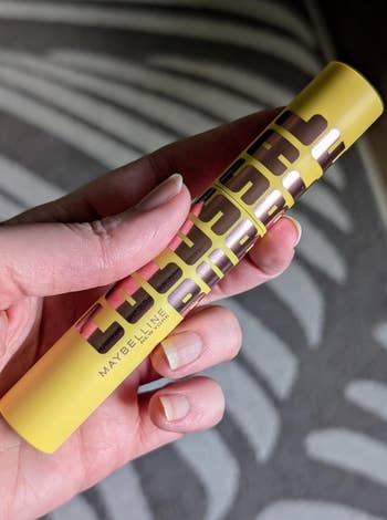A idiosyncratic   holds a Maybelline Lash Mascara conduit  with bold lettering, placed against a subtle striped background