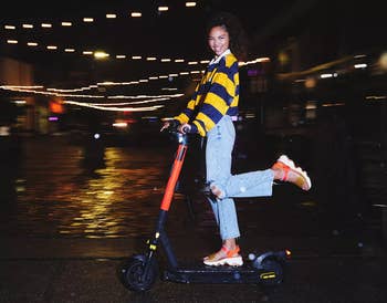 Person riding a scooter astatine  nighttime  wearing a striped sweater, ripped jeans, and sneakers, smiling joyfully against a backdrop of metropolis  lights